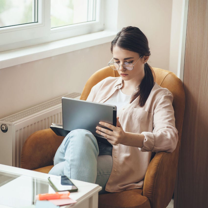 Konzentrierte junge Frau mit Brille, die im Sessel sitzend am Laptop arbeitet