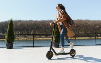 Frau in Mantel fährt auf E-Scooter an einem Kanal entlang