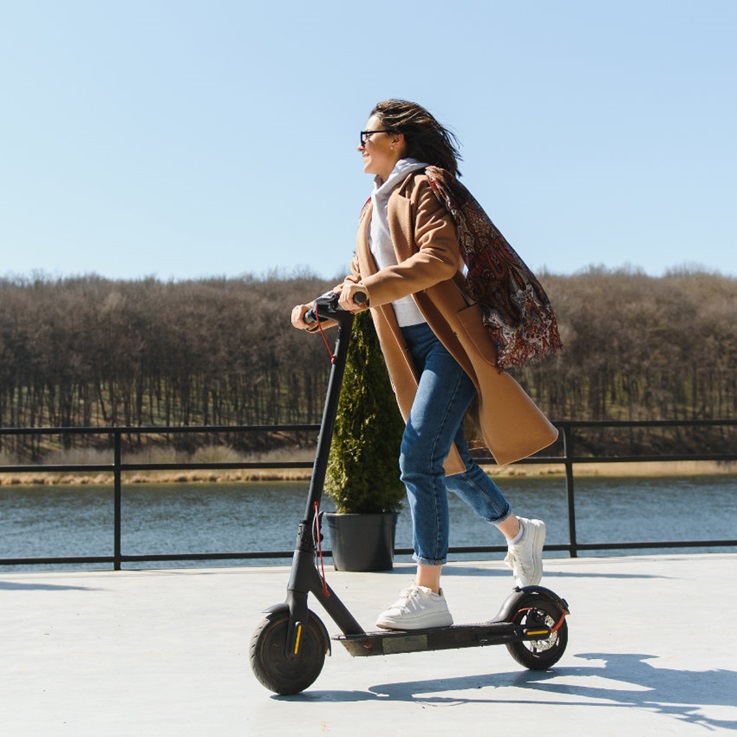 Frau in Mantel fährt auf E-Scooter an einem Kanal entlang