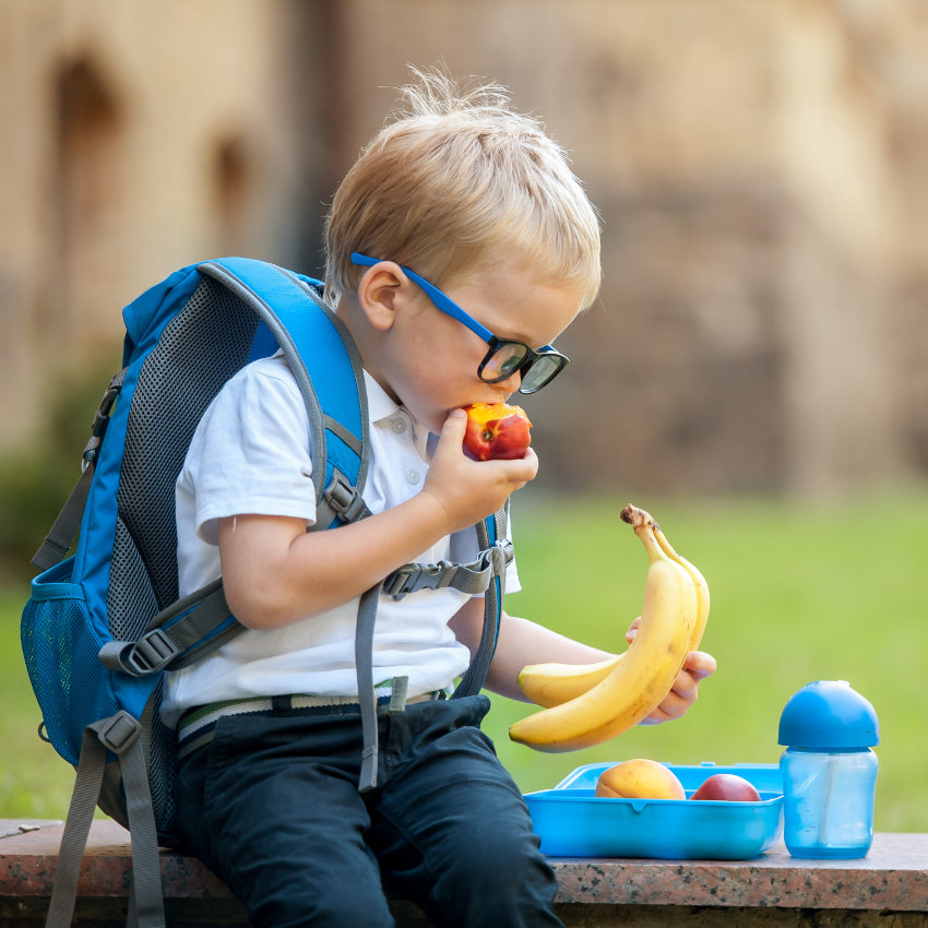 Kleiner Junge mit Rucksack und Brotdose isst Obst