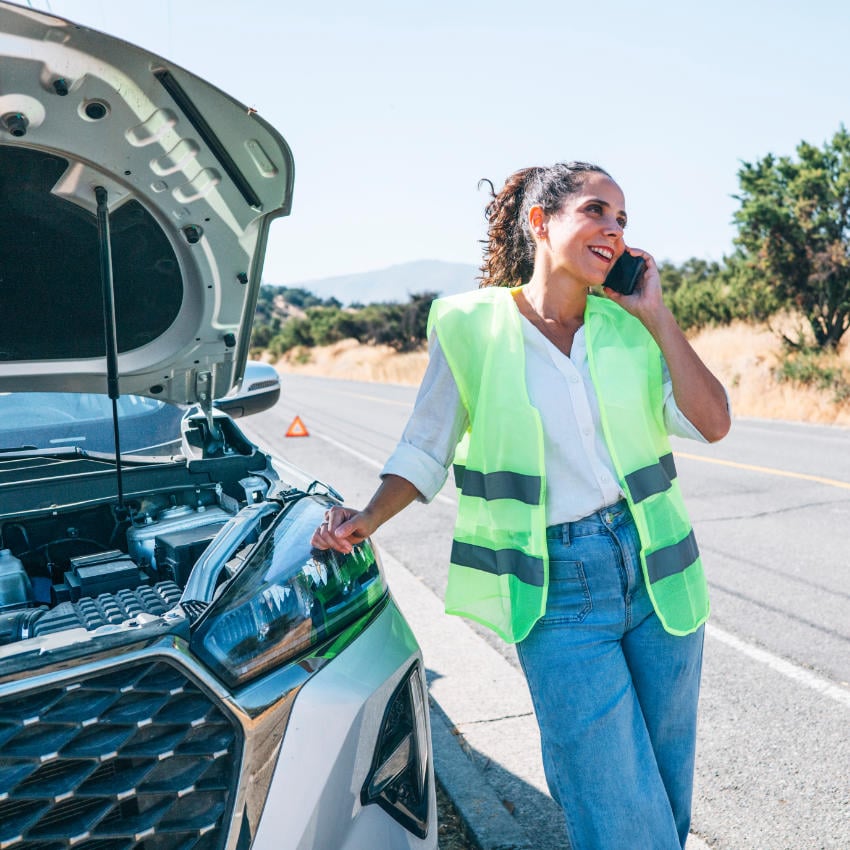 mallorca-police_850x850_2022_02 Frau in Warnweste steht an einer ländlichen Straße neben ihrem liegengebliebenen Auto, telefoniert mit dem Mobiltelefon, während die Motorhaube offen ist und ein Warndreieck hinter dem Fahrzeug aufgestellt wurde