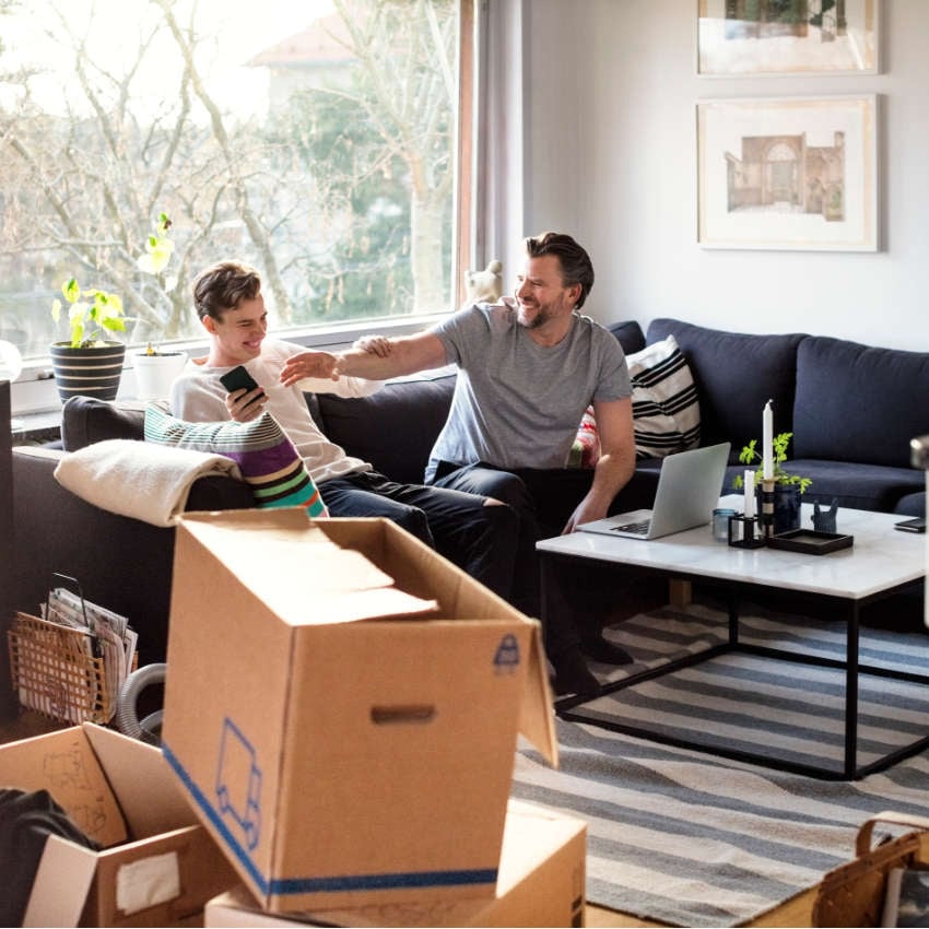 GettyImages-681890213 Vater und Sohn, die zusammen in einem Wohnzimmer mit Umzugskartons auf einem Sofa sitzen und lachen