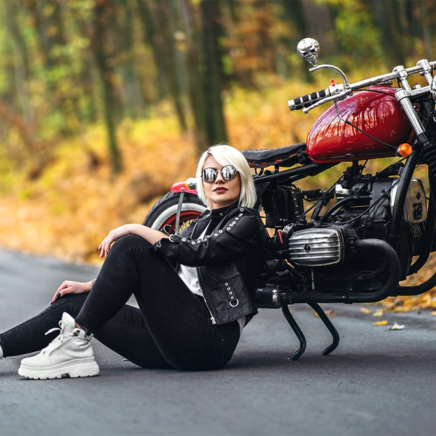 Frau mit Sonnenbrille sitzt vor einem roten Motorrad auf einer Straße im herbstlichen Wald