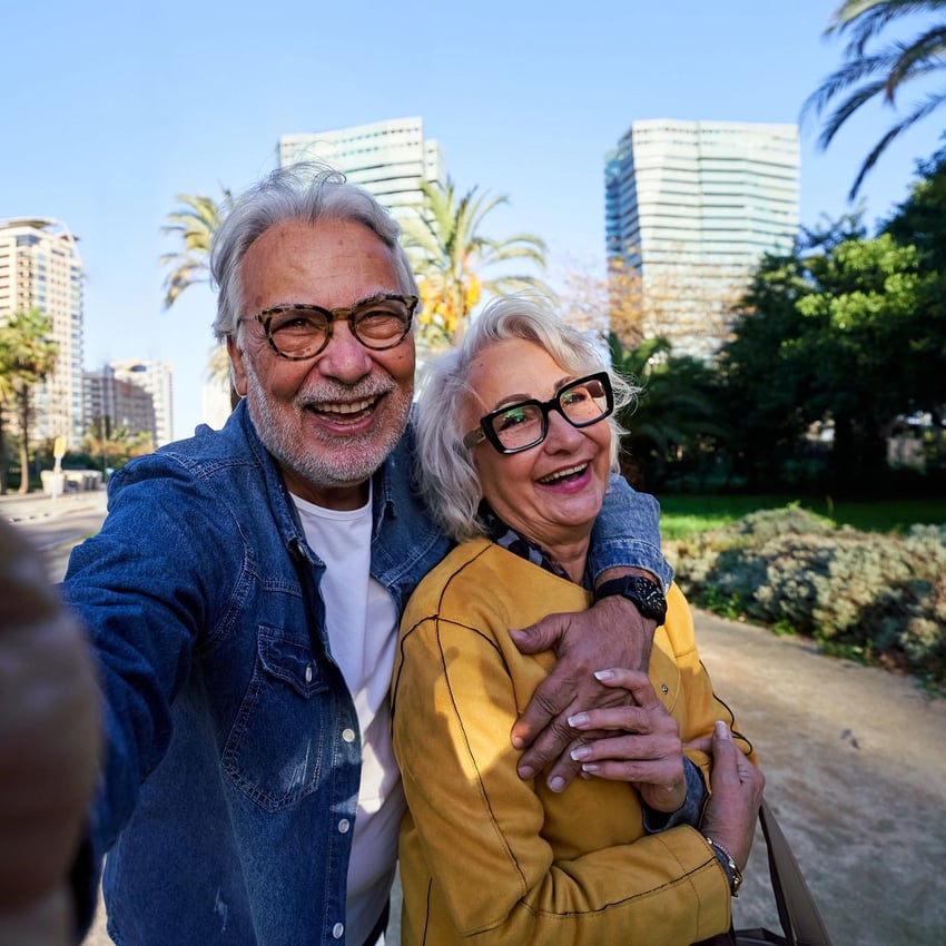 Älteres Paar nimmt gemeinsam ein Selfie in einem sonnigen Stadtpark mit modernen Gebäuden im Hintergrund auf