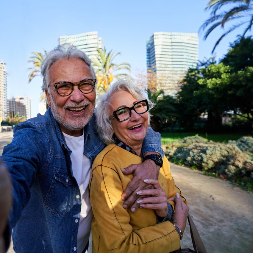 Älteres Paar nimmt gemeinsam ein Selfie in einem sonnigen Stadtpark mit modernen Gebäuden im Hintergrund auf