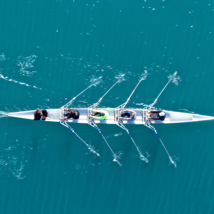 Ruderboot mit vier Ruderern und einer Steuerperson, aufgenommen aus der Vogelperspektive auf ruhigem, türkisblauem Wasser