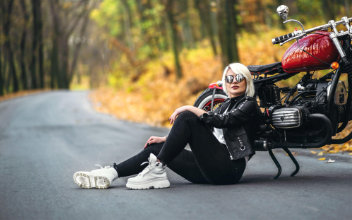 Frau mit Sonnenbrille sitzt vor einem roten Motorrad auf einer Straße im herbstlichen Wald