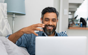 Mann sitzt auf Sofa und telefoniert mit Laptop auf dem Schoss