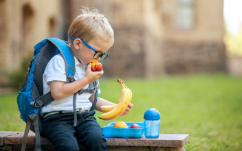 Kleiner Junge mit Rucksack und Brotdose isst Obst