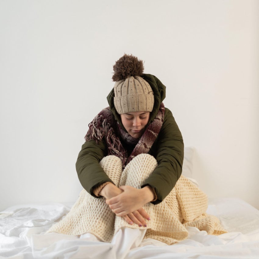 Frau sitzt frierend mit Mütze und Winterjacke in ihrer Wohnung Frierende Frau